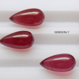 Op de afbeelding: Vijf peervormige rode edelstenen op een wit oppervlak. De edelstenen zijn glad en glanzend. GEMSONLY is gedrukt op het oppervlak.