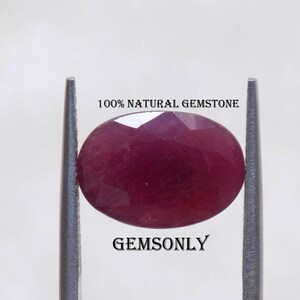 May include: A close-up of a deep red oval gemstone held in place by two metal tweezers. The gemstone is smooth and shiny, with a slight sparkle. The text "100% NATURAL GEMSTONE" and "GEMSONLY" are visible above and below the gemstone.