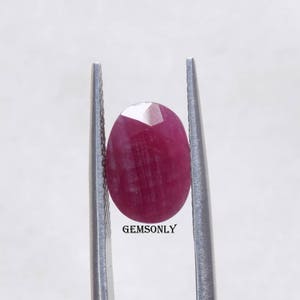 May include: A close-up of a red oval gemstone held in tweezers. The gemstone is faceted and has a slight shimmer.