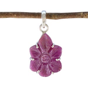May include: A carved, flower-shaped pendant in shades of pink and purple, suspended from a silver bail. The pendant is detailed with a central design and silver accents, hanging from a thin, brown branch against a white background.