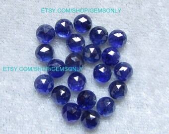 May include: A collection of 20 faceted sapphire gemstones, each with a deep blue color. The gemstones are round and have a polished finish. They are arranged in a loose pile on a white surface.