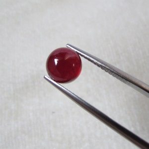 May include: A round, polished, deep red gemstone held by a pair of silver tweezers. The gemstone has a smooth, reflective surface, and the tweezers are positioned at an angle against a soft, white background.