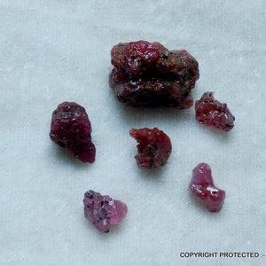 Puede incluir: Seis piedras preciosas de rubí en bruto, de color rosa rojizo, de diferentes tamaños y formas. Las piedras preciosas están sobre una superficie blanca.