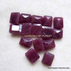 May include: A collection of 12 purple gemstone cabochons, cut in a rectangular shape. The gemstones are arranged in a loose pile on a white surface.  GEMSONLY BY PUNEET is printed on the surface.