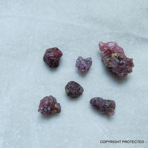 May include: A collection of six pink and purple rough ruby crystals on a white surface.