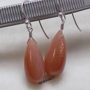 May include: A pair of silver dangle earrings with teardrop-shaped sunstone gemstones. The stones are a light orange color with a shimmery, iridescent finish.