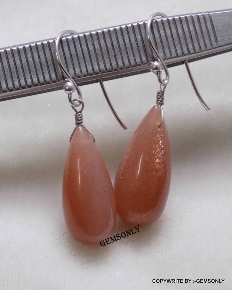 May include: A pair of silver dangle earrings with teardrop-shaped sunstone gemstones. The stones are a light orange color with a shimmery, iridescent finish.