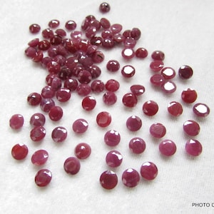 Puede incluir: Una colección de aproximadamente 50 piedras preciosas de rubí facetadas sueltas. Los rubíes son de color rojo intenso y tienen una forma redonda. Los rubíes están dispersos sobre una superficie blanca.