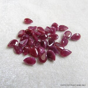 May include: A collection of pear-shaped ruby gemstones, ranging in size and shade of red. The gemstones are arranged in a loose pile on a white surface.