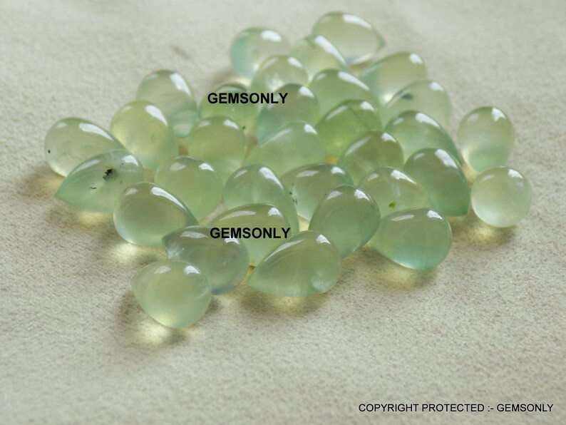 May include: A collection of translucent, pale green gemstones in various shapes, including teardrops and rounded forms. The gems are scattered across a neutral-toned surface, with the text "GEMSONLY" visible in the image.
