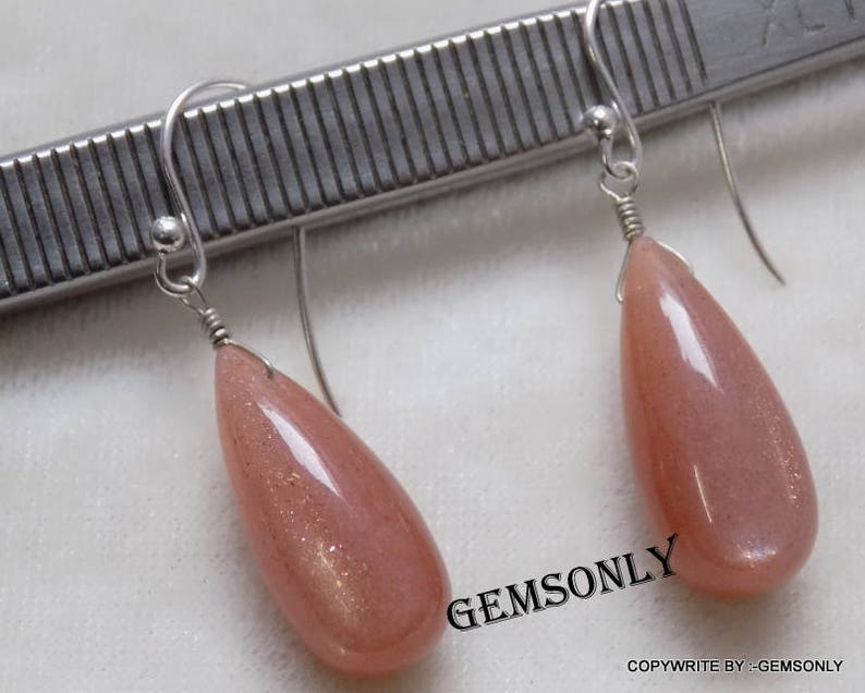 May include: A pair of silver earrings with teardrop-shaped sunstone gemstones. The stones are a light peach color with gold flecks.