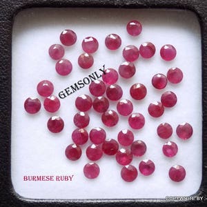 Puede incluir: Una colección de 30 rubíes birmanos sueltos, redondos y rojos. Los rubíes se muestran en un recipiente de plástico blanco cuadrado con un borde negro. El recipiente está etiquetado con "BURMESE RUBY" en texto negro.