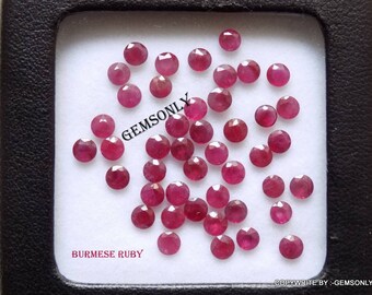 May include: A collection of 30 loose, round, red Burmese rubies. The rubies are displayed in a white, square, plastic container with a black border. The container is labeled "BURMESE RUBY" in black text.
