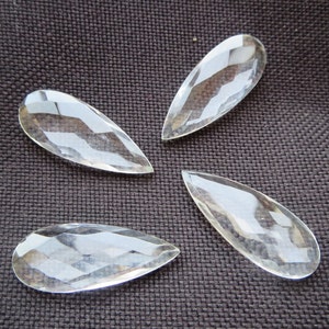 May include: Four clear glass teardrop-shaped crystal prisms, each with a faceted design, are arranged in a cross pattern on a brown fabric surface.