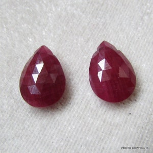 May include: Two pear-shaped ruby gemstones with a faceted cut. The rubies are a deep red colour and have a glossy finish.