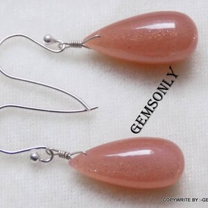 May include: A pair of silver earrings with teardrop-shaped sunstone stones. The stones are a light peach color with a shimmery finish.
