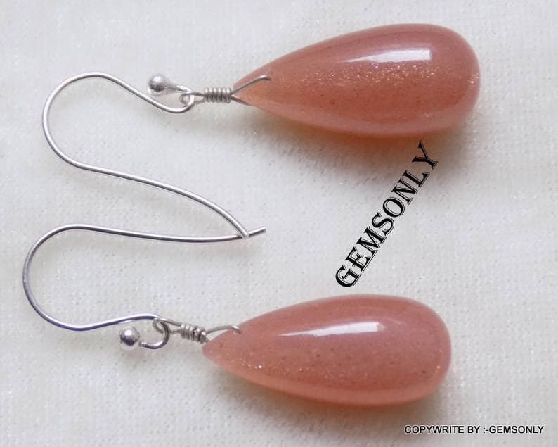 May include: A pair of silver earrings with teardrop-shaped sunstone stones. The stones are a light peach color with a shimmery finish.