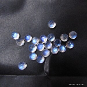 Pode incluir: Uma coleção de 17 cabochões redondos de pedra da lua iridescente em vários tamanhos, com um brilho azul-branco, exibidos em uma superfície preta.