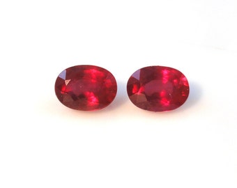 May include: Two oval-cut, deep red gemstones are displayed against a white background. The gems have a faceted surface, catching the light and displaying a vibrant, rich color. The stones are similar in size and shape.
