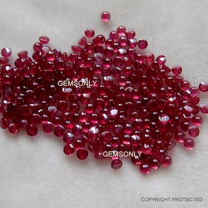 Può includere: Un grande gruppo di piccole pietre preziose di rubino rotonde e rosse. Le pietre preziose sono sparse su una superficie bianca.