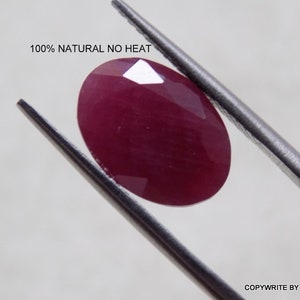 May include: A deep red oval-shaped gemstone, 100% natural and untreated, held by tweezers against a white background. The text "100% NATURAL NO HEAT" is visible above the gemstone.