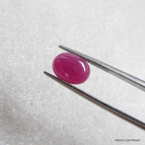 May include: A vibrant pink oval-shaped gemstone held by tweezers. The gemstone is smooth and polished, with a glossy finish.