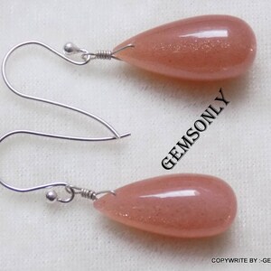 May include: A pair of silver earrings with teardrop-shaped peach sunstone gemstones. The earrings have a simple design with a small silver ball at the top of the hook.