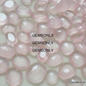 Pu&ograve; includere: Una collezione di pietre preziose di quarzo rosa pallido, ciascuna con una forma sfaccettata irregolare. Le pietre sono sparse su una superficie bianca.