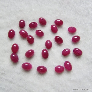 May include: A collection of twenty oval-shaped, faceted ruby gemstones. The rubies are a deep red color and are scattered on a white surface.