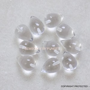 May include: Ten clear teardrop-shaped crystal beads with a hole drilled through the top. The beads are arranged in a circle on a white surface.