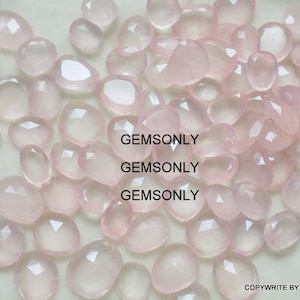 Pu&ograve; includere: Una collezione di pietre preziose di quarzo rosa pallido, ciascuna con una forma sfaccettata irregolare. Le pietre sono sparse su una superficie bianca. GEMSONLY &egrave; stampato tre volte sull'immagine.