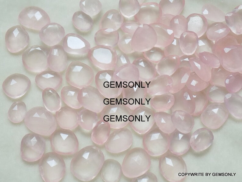 Pu&ograve; includere: Una collezione di pietre preziose di quarzo rosa pallido, ciascuna con una forma sfaccettata irregolare. Le pietre sono sparse su una superficie bianca. GEMSONLY &egrave; stampato tre volte sull'immagine.