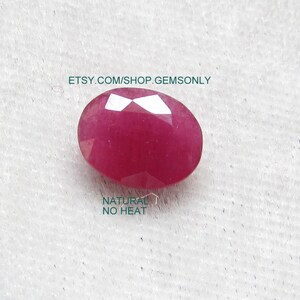 May include: A vibrant pink oval-shaped ruby gemstone with a faceted cut. The gemstone is set against a white background. The text "NATURAL NO HEAT" is visible below the gemstone.