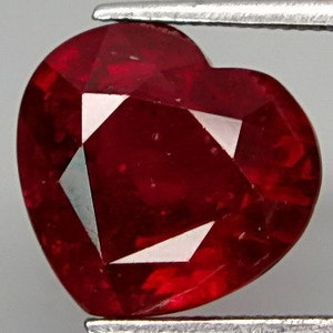 May include: A heart-shaped ruby gemstone with a deep red color. The gemstone is faceted and has a polished finish.