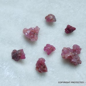 Puede incluir: Una colección de gemas de rubí en bruto sin cortar. Los rubíes varían en tamaño y forma, mostrando un tono rosa intenso a rosa rojizo. Las gemas tienen una apariencia natural, sin pulir, con estructuras cristalinas visibles.