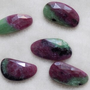 May include: Five oval-shaped green and pink gemstones with faceted edges. The stones are arranged on a white fabric background.