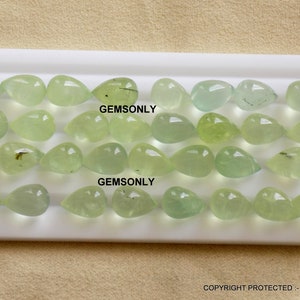 May include: A collection of light green, teardrop-shaped gemstones arranged on a white surface. The gemstones vary in shade, from pale to vibrant green, and are translucent. The text "GEMSONLY" is visible on the surface. The image is well-lit, showcasing the gems' clarity.
