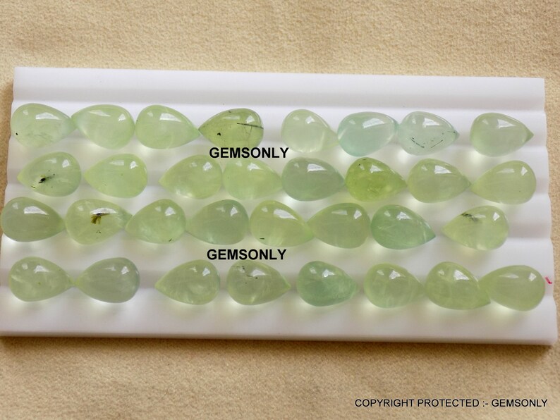 May include: A collection of light green, teardrop-shaped gemstones arranged on a white surface. The gemstones vary in shade, from pale to vibrant green, and are translucent. The text "GEMSONLY" is visible on the surface. The image is well-lit, showcasing the gems' clarity.