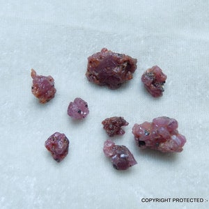 May include: A collection of eight pink ruby rough stones. The stones are various sizes and shapes, with some having a more crystalline appearance.