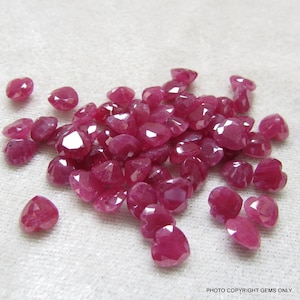 Op de afbeelding: Een stapel hartvormige robijn edelstenen, met een dieprode kleur en gefacetteerde oppervlakken. De edelstenen zijn verspreid over een wit oppervlak.