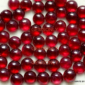 Puede incluir: Una colección de cabujones redondos de piedras preciosas rojas. Los cabujones son lisos y pulidos, con un acabado de alto brillo. Los cabujones son todos de tamaño y forma aproximadamente iguales. El texto "GEMSONLY" aparece tres veces en la imagen.