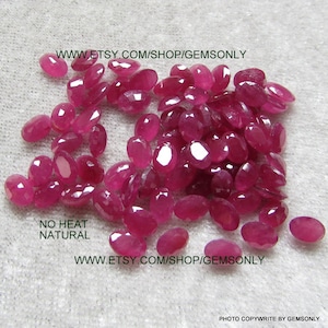May include: A collection of oval-cut, natural, no-heat rubies in varying sizes. The gems are a deep, vibrant red color and are scattered across a light-colored surface. The image includes the text "NO HEAT NATURAL" and a website address.