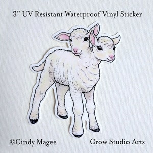 May include: A 3" UV resistant waterproof vinyl sticker featuring a cartoon illustration of a two-headed white lamb. The sticker is printed with the text "Cindy Magee" and "Crow Studio Arts".