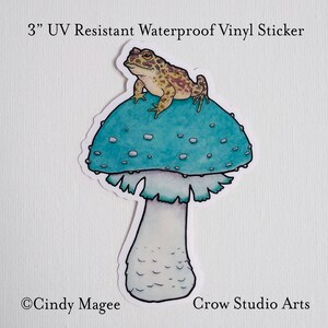 May include: A 3" UV resistant waterproof vinyl sticker featuring a cartoon toad sitting on a blue and white mushroom. The sticker says "Crow Studio Arts" at the bottom.