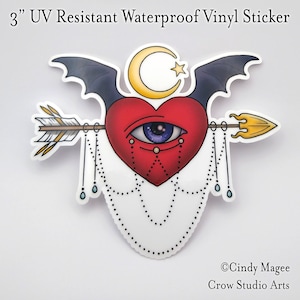 May include: A 3 inch vinyl sticker featuring a red heart with a blue eye pierced by an arrow. The heart is adorned with a chain of small circles and teardrop shapes. A crescent moon and a star are above the heart, and bat wings are behind the moon and star.