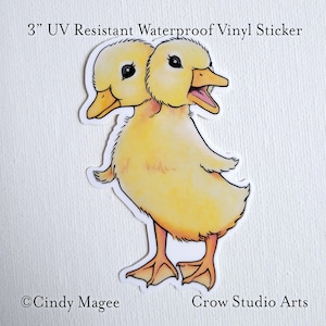 May include: A 3" UV resistant waterproof vinyl sticker featuring a cartoon illustration of a two-headed yellow duckling with orange feet. The sticker is on a white background with the text "Cindy Magee" and "Crow Studio Arts" at the bottom.