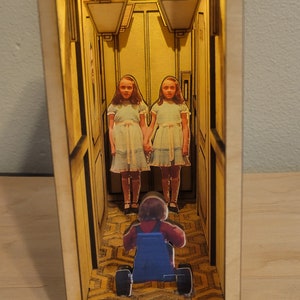 Puede incluir: Un diorama de madera que representa un pasillo con dos figuras con vestidos blancos de pie al final del pasillo. Una figura con un mono azul est&aacute; montando un triciclo de juguete hacia las figuras.