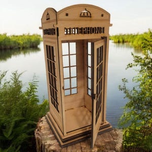 May include: A wooden model of a classic red telephone booth with an open door. The booth is made of laser-cut wood and has a detailed design. The booth is sitting on a tree stump in front of a body of water.