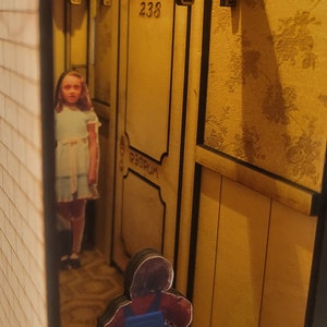 Puede incluir: Un diorama en miniatura de un pasillo con una puerta etiquetada como "238" y la palabra "Dormitorio" debajo. Una ni&ntilde;a con un vestido azul se encuentra en el pasillo, y una figura con una camisa roja y un mono azul monta un coche de juguete en un suelo estampado.