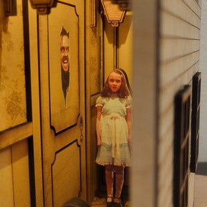 Puede incluir: Un modelo de madera de una casa con un interior iluminado. La casa tiene una puerta con un recorte de la cara de un hombre y una ventana con un recorte de una ni&ntilde;a con un vestido azul. Hay una figura de una persona con un mono azul sentada frente a la casa.